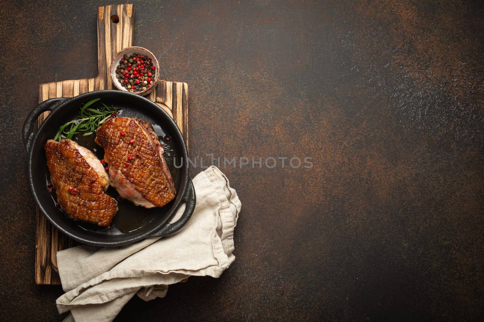 Two roasted duck breast fillets with crispy skin, with pepper and rosemary, top view in black cast iron pan with knife, dark brown concrete rustic background, space for text by its_al_dente