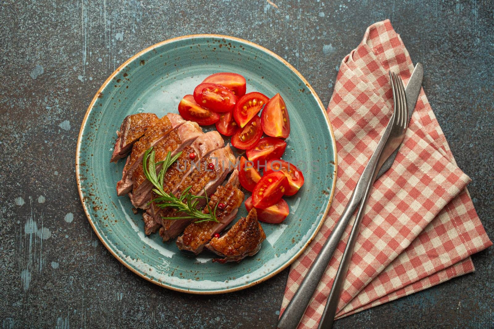 Delicious roasted sliced duck breast fillet with golden crispy skin, with pepper and rosemary, top view on ceramic blue plate served with cherry tomatoes salad, rustic concrete rustic background by its_al_dente