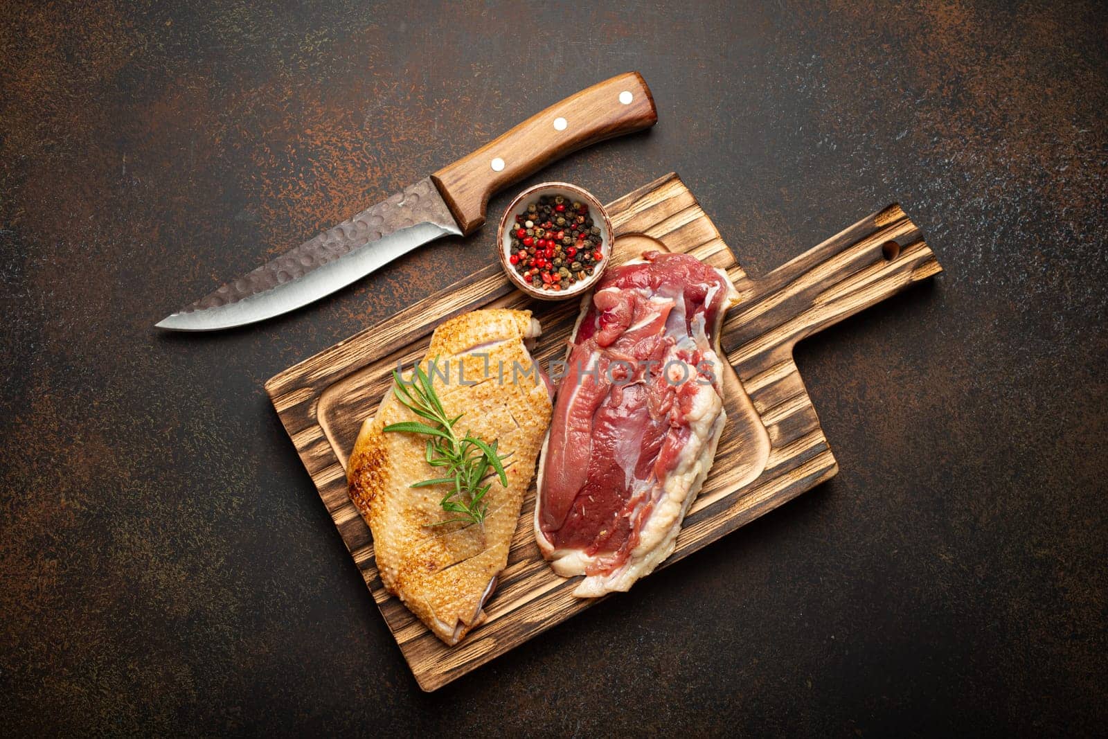 Two raw uncooked duck breast fillets with skin, seasoned with salt, pepper, rosemary top view on wooden cutting board with knife, dark brown concrete rustic background by its_al_dente