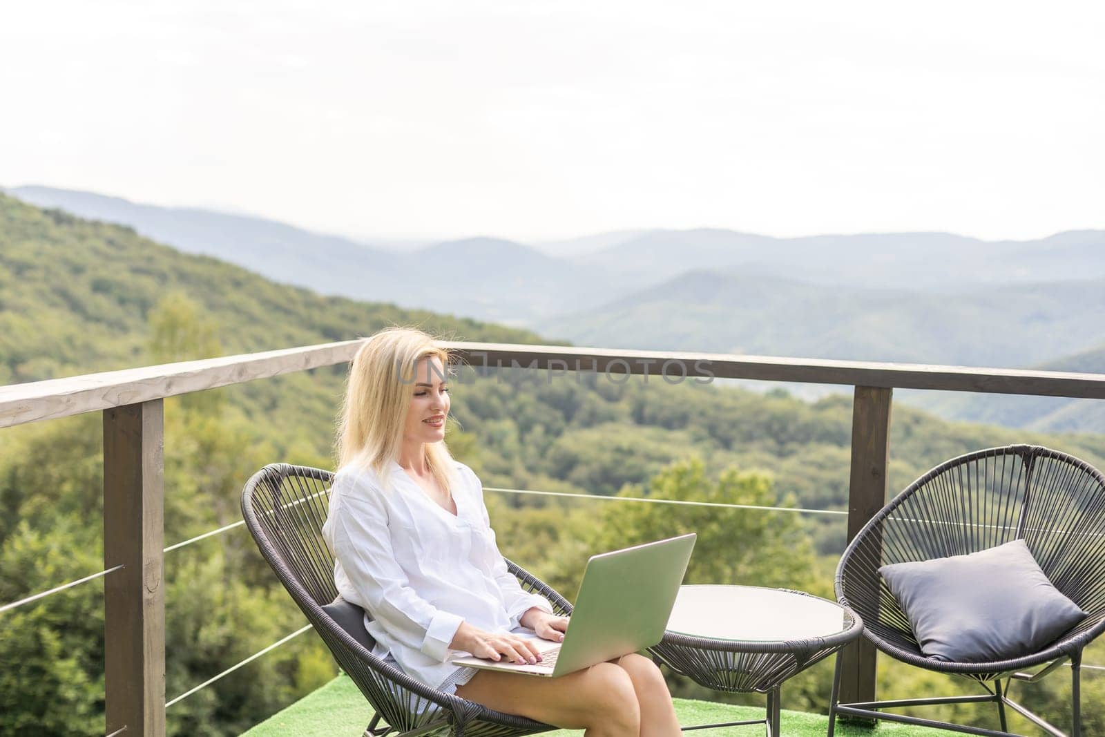 A young woman, freelancer is working on a laptop remotely on a balcony in the open fresh air near the mountains in warm sunny weather