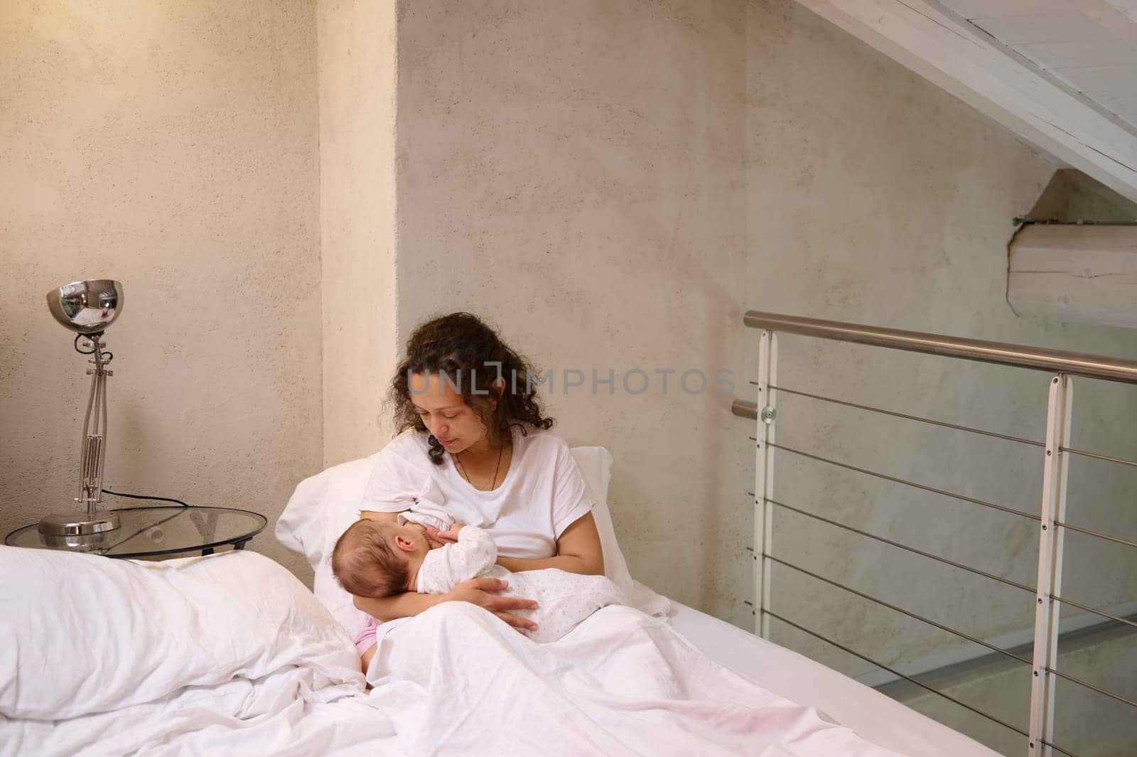 Young curly haired multi ethnic woman, loving caring mother holding and breastfeeding her newborn baby, sitting on the bed in white bedchamber interior. Family relationships. Maternity lifestyle
