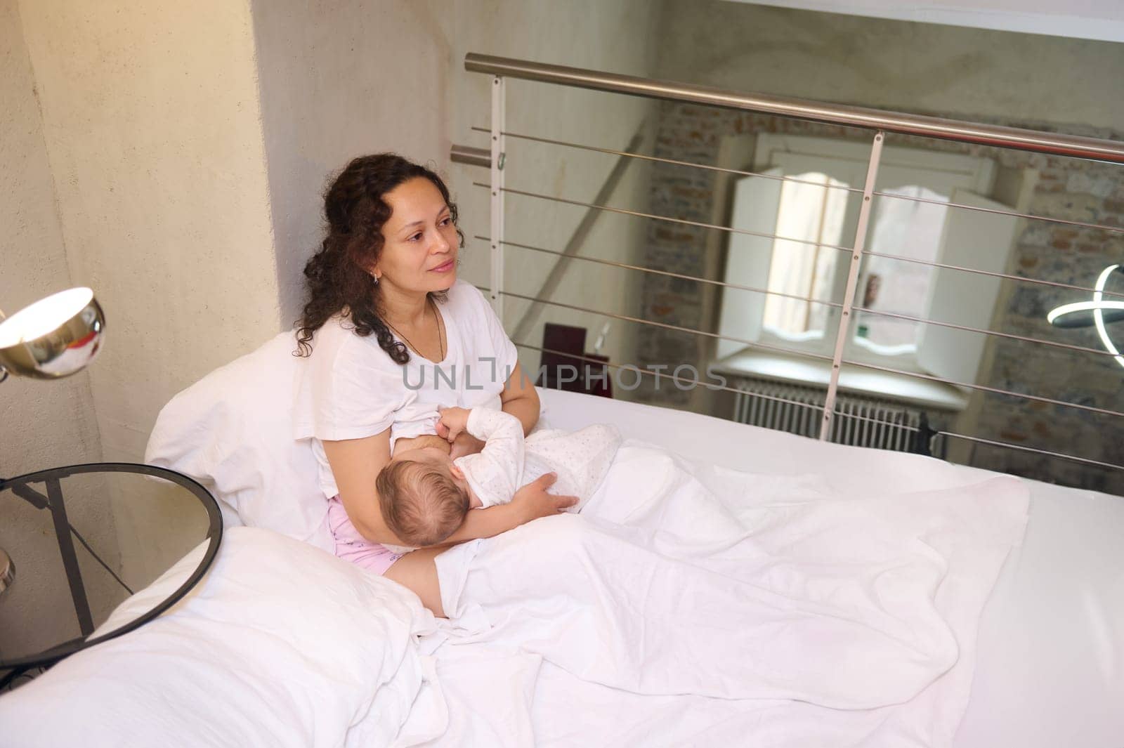View from above of loving caring mother and her cute newborn baby. Happy mother sitting on the bed in cozy home interior, smiles looking at her baby boy while breastfeeding him. Tenderness. Love. Care