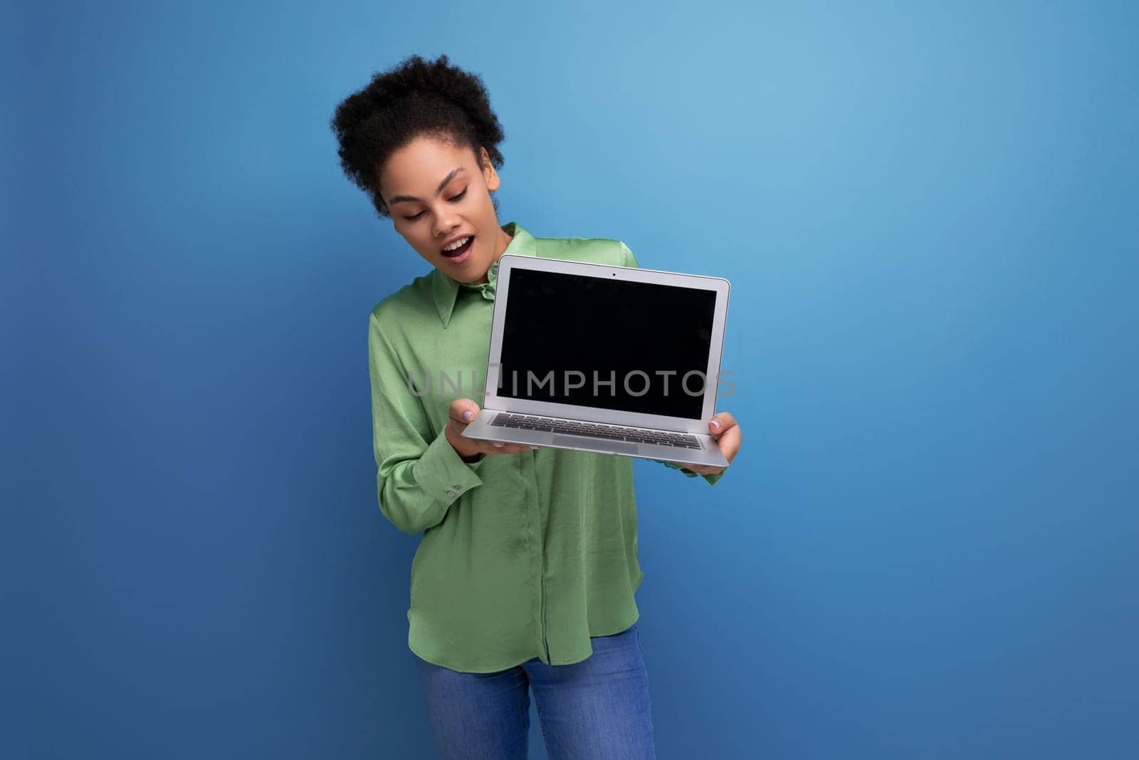 young pretty female model with curly black hair dressed in a green blouse and jeans holds a laptop screen forward with a mock up for advertising by TRMK