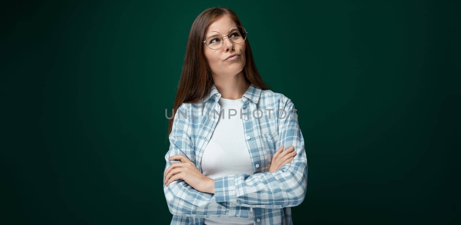 Young European woman dressed in a plaid shirt and jeans thinking on a dark green background by TRMK