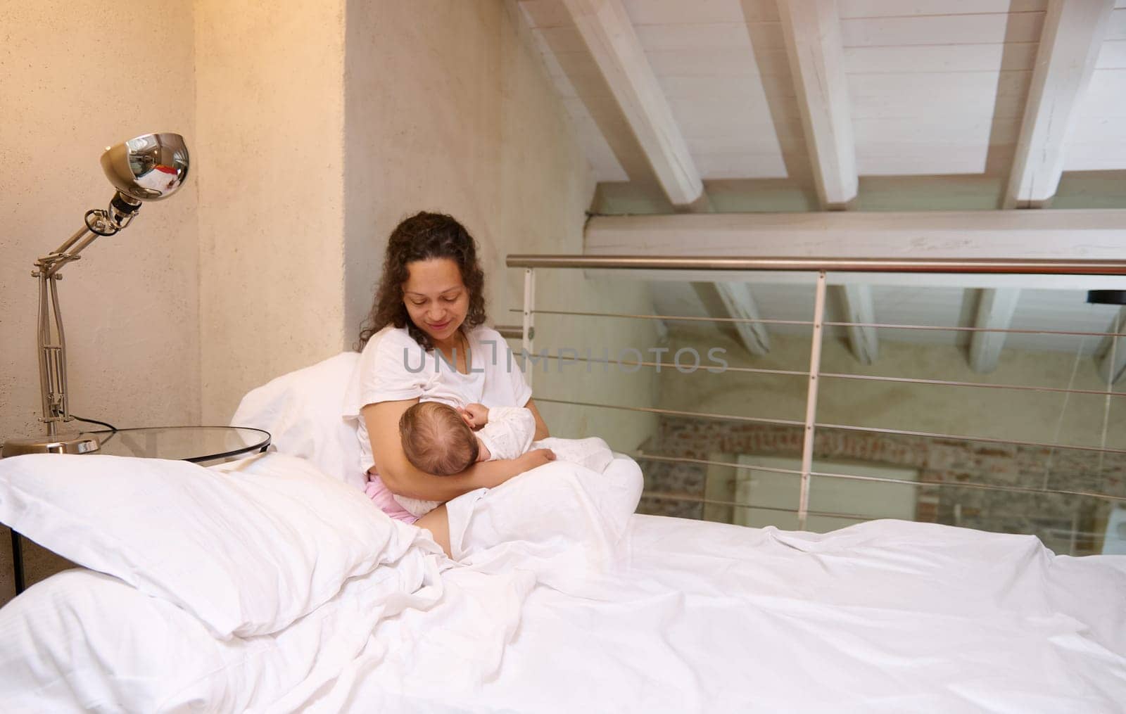 Portrait of a loving caring mother and her cute newborn baby. Happy mother sitting on the bed in cozy home interior, smiles looking at her baby boy while breastfeeding him. Tenderness. Love. Care