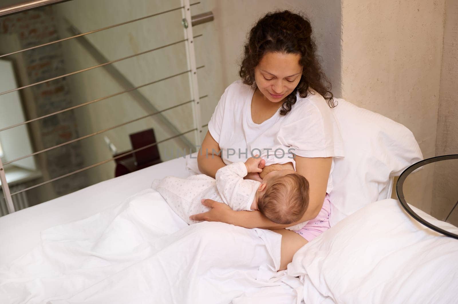Confident Latin American young beautiful woman, loving caring mother enjoys her maternity leave lifestyle, holds her newborn baby in her arms and breastfeeds him while wakes up in the morning