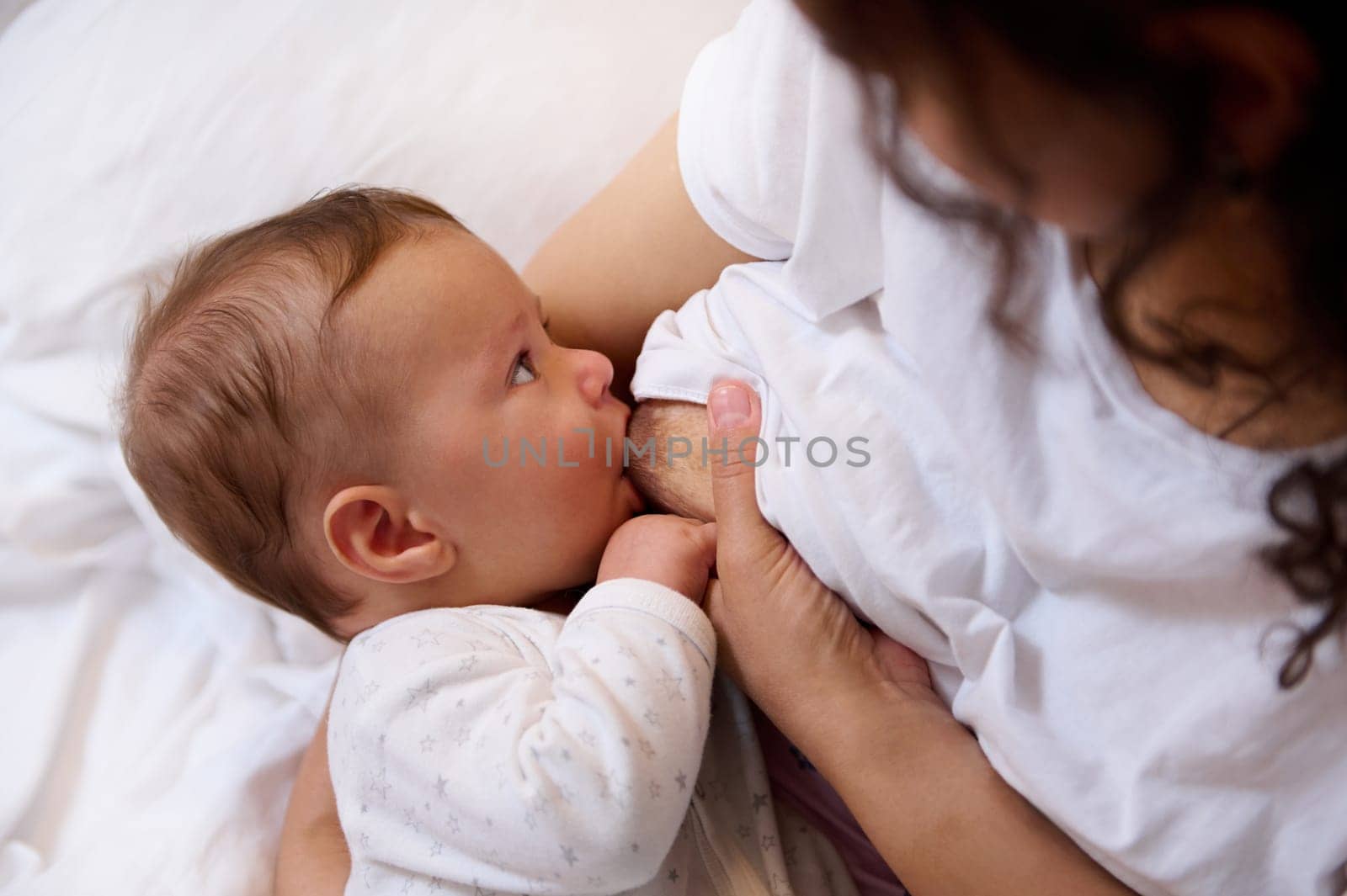 Close-up of Caucasian adorable baby boy dressed in white clothes, suckling the healthy milk from the breast of his mother. Breastfeeding as one way to reduce the risk and prophylaxis of breast cancer
