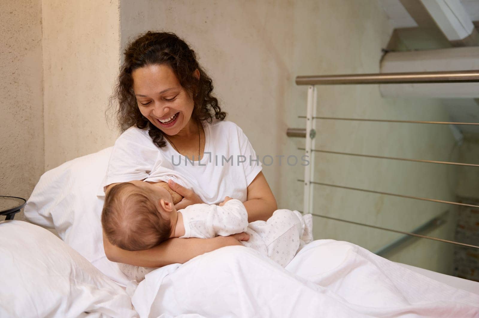 Happy mother smiling breastfeeding newborn baby, sitting on the bed in white bedchamber, expressing happy positive emotions, feeling emotional connection with her beloved infant. Maternity lifestyle