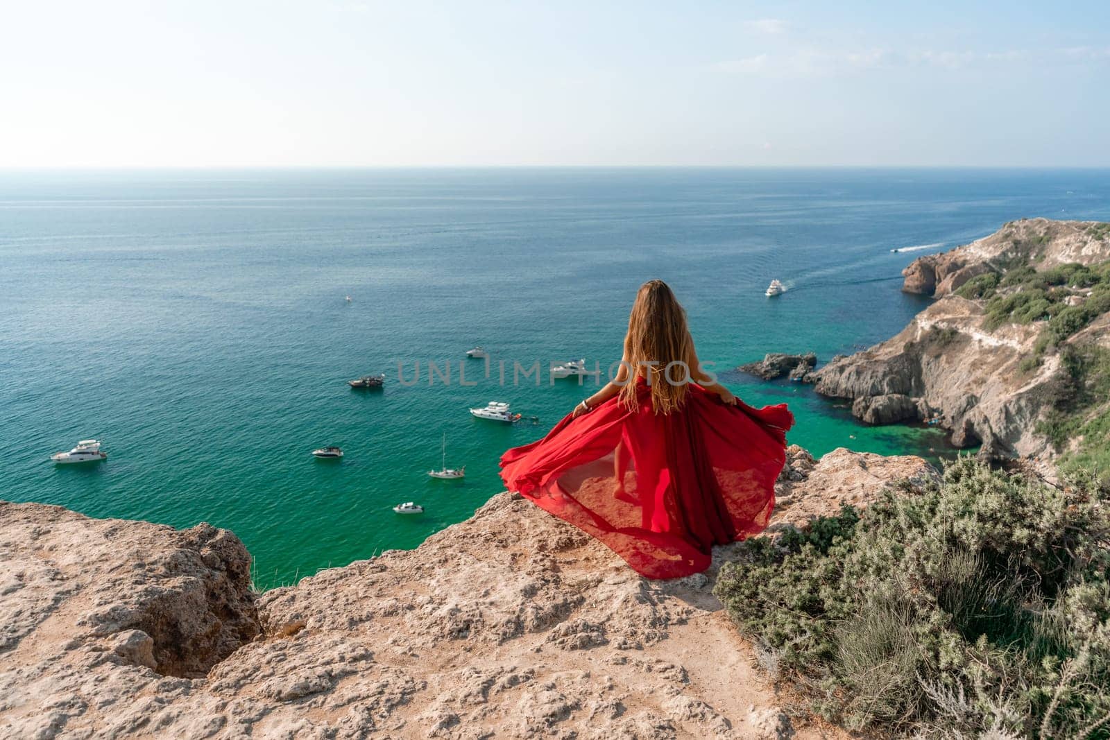 Woman sea red dress yachts. A beautiful woman in a red dress poses on a cliff overlooking the sea on a sunny day. Boats and yachts dot the background