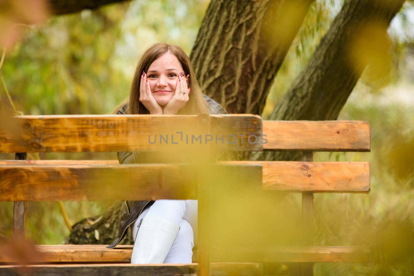 A young beautiful girl sits on a bench in an autumn park and looks cheerfully into the frame a