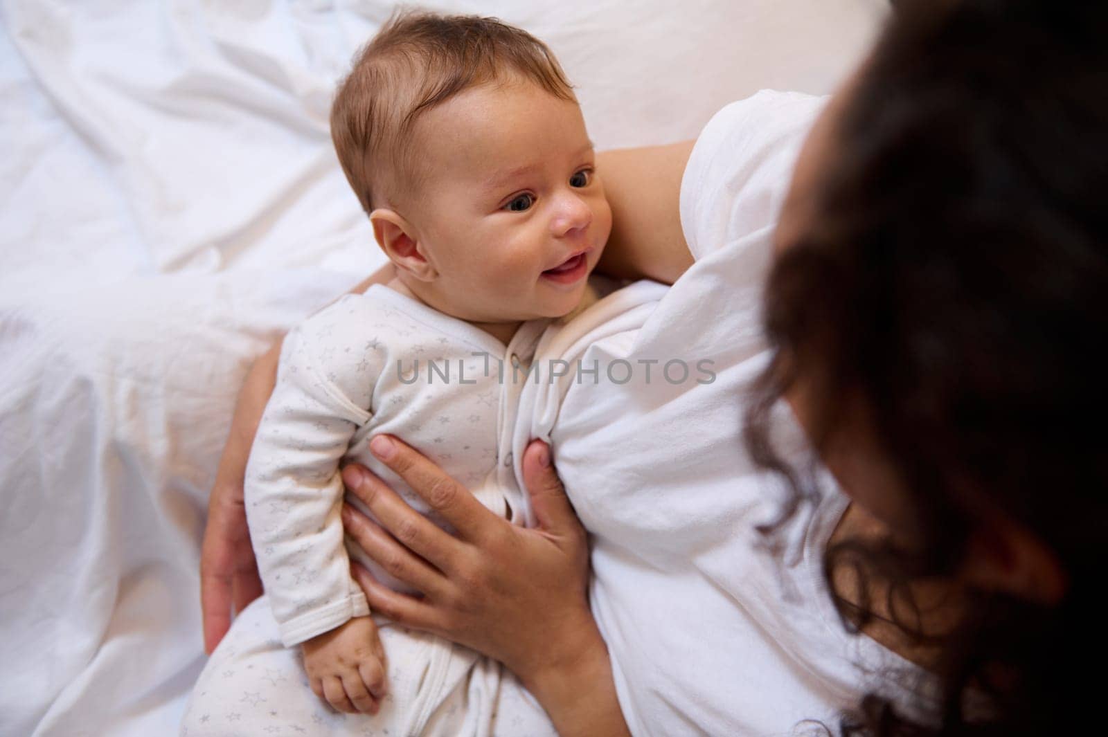 View from above of Caucasian adorable cute newborn baby boy smiles cutely, looking at his loving caring mother hugging and holding him in her arms. Maternity leave and infancy. Tenderness. Love. Care