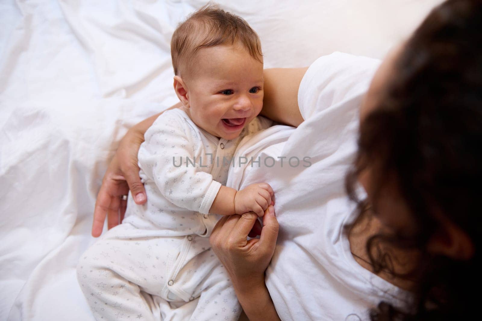 Authentic lifestyle portrait of a multi ethnic happy woman mother cuddling her baby boy, sitting together on the bed in bedchamber interior. People. Maternity leave lifestyle. Copy advertising space