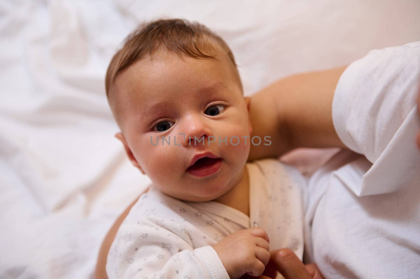 Close-up portrait of Caucasian adorable cute lovely blue eyes newborn baby boy in white clothes, looking at camera while his mom cuddles him. Children. Infancy and babyhood. Maternity leave lifestyle