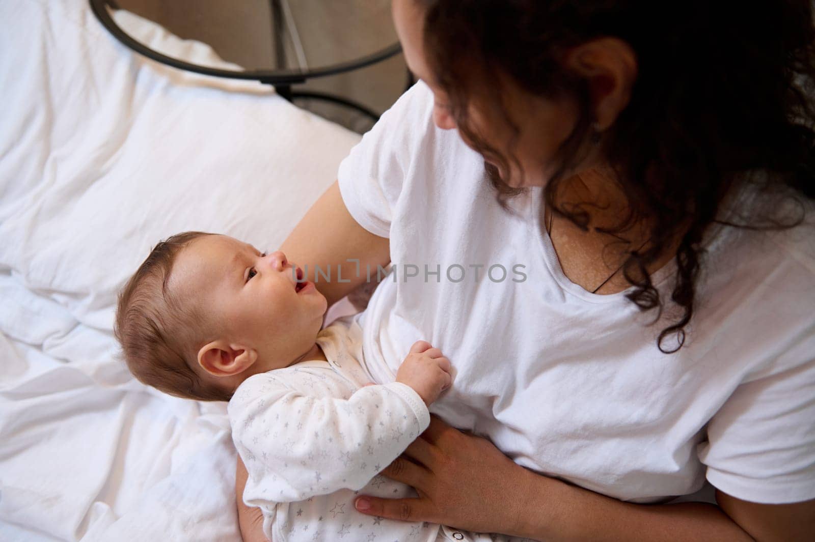 Authentic portrait of curly brunette young adult woman, happy loving mother smiling, expressing positive emotions while breastfeeding and cuddling her newborn baby. People. Maternity leave lifestyle.