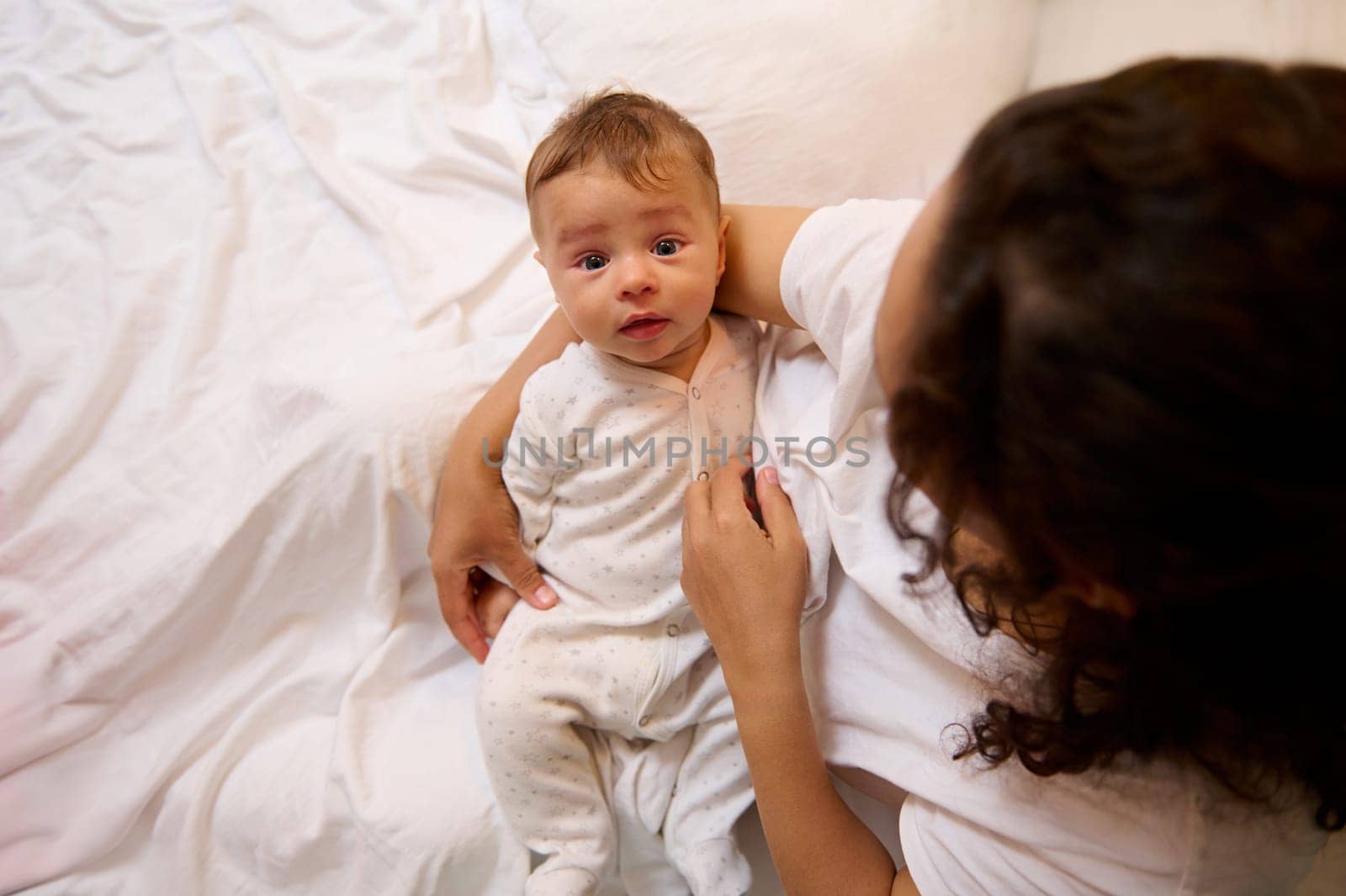 Portrait of authentic little baby boy in the hands of his mother, sitting together on the bed in cozy bedroom interior. Maternity leave lifestyle, infancy, babyhood. Motherhood. Single parenting