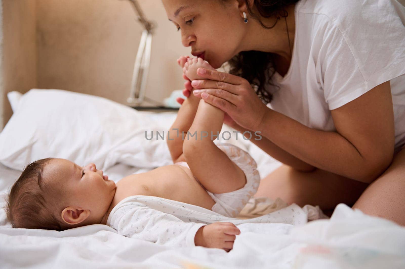 Beautiful Latin American woman, young loving caring mother kissing her newborn baby boy feet, expressing positive emotions of happy moments spending together and maternity leave and babyhood concept
