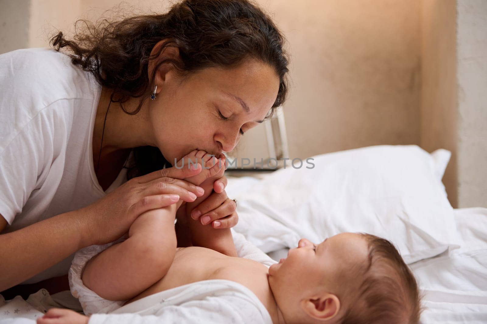 Closeup young loving caring mom gently kisses little feet and tiny toes of her newborn baby boy, enjoying spending tender moments with her beloved kid. Mother and child connection. Baby care concept