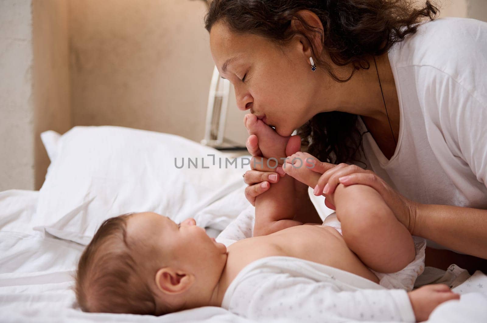 Adorable baby boy of four month old smiles looking at his mom, while she kisses his little feet and toes, enjoying happy moments together. Delightful mother enjoys wonderful moments with her child