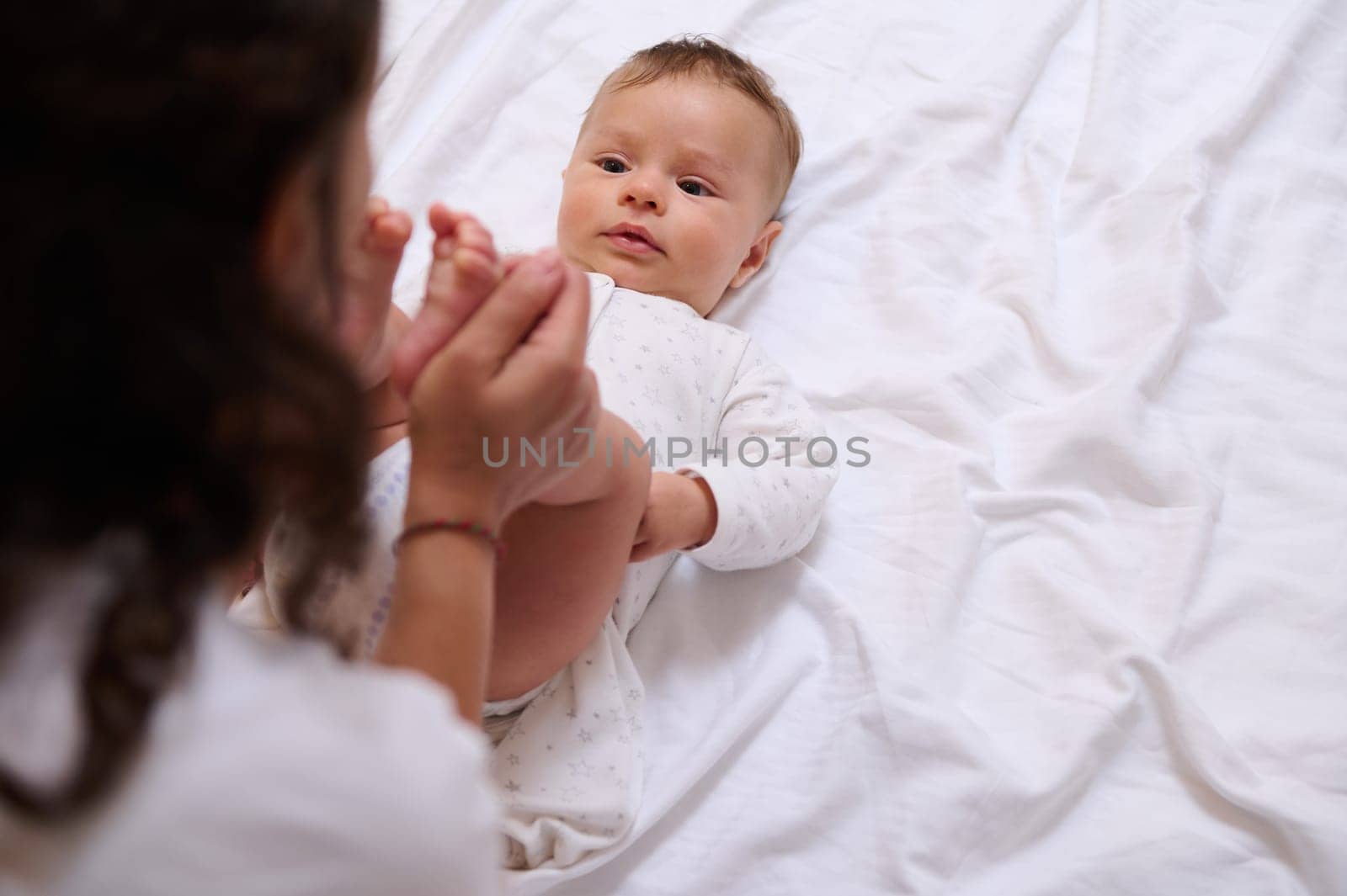 Overhead view of a multi ethnic loving mom kissing little feet and tiny toes of her adorable beloved baby boy. Baby care, infancy, babyhood, maternity leave lifestyle concept. Copy advertising space