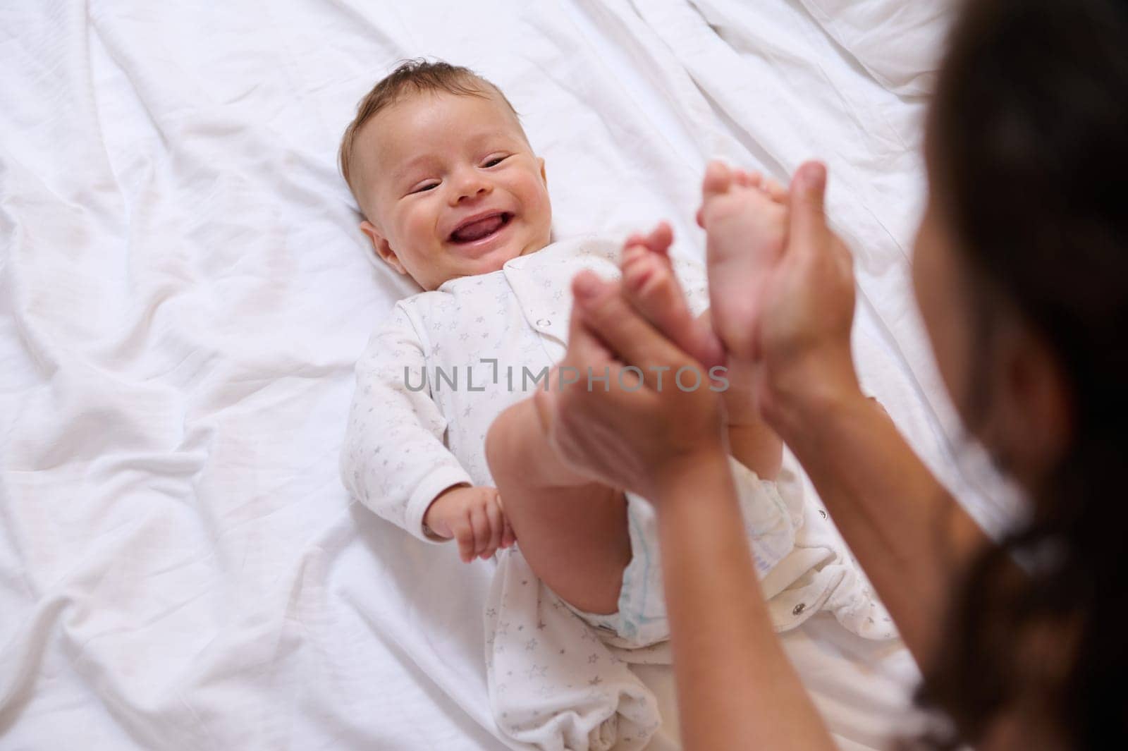 Caucasian beautiful newborn baby boy, cutely smiles lying on the bed and looking at his loving caring mother, while she strokes and kisses his little feet and tiny toes. Family. Infancy and motherhood