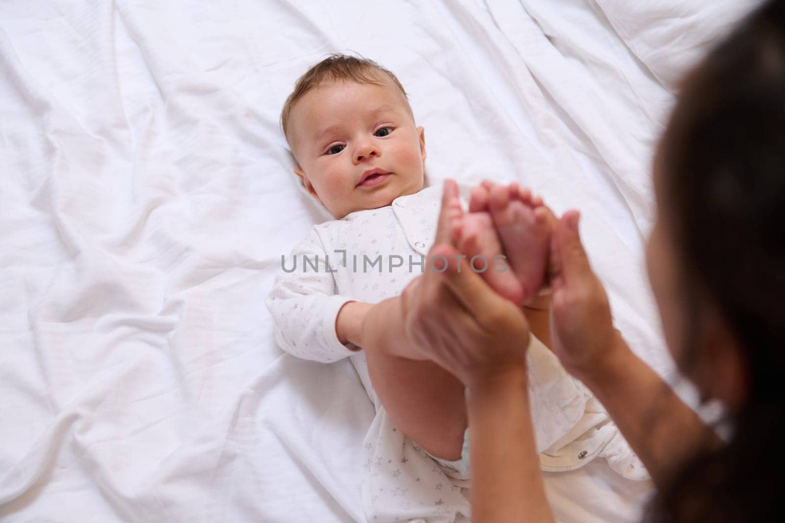 Adorable newborn baby looks at camera while his loving caring mom strokes and kisses his little feet and tiny toes by artgf