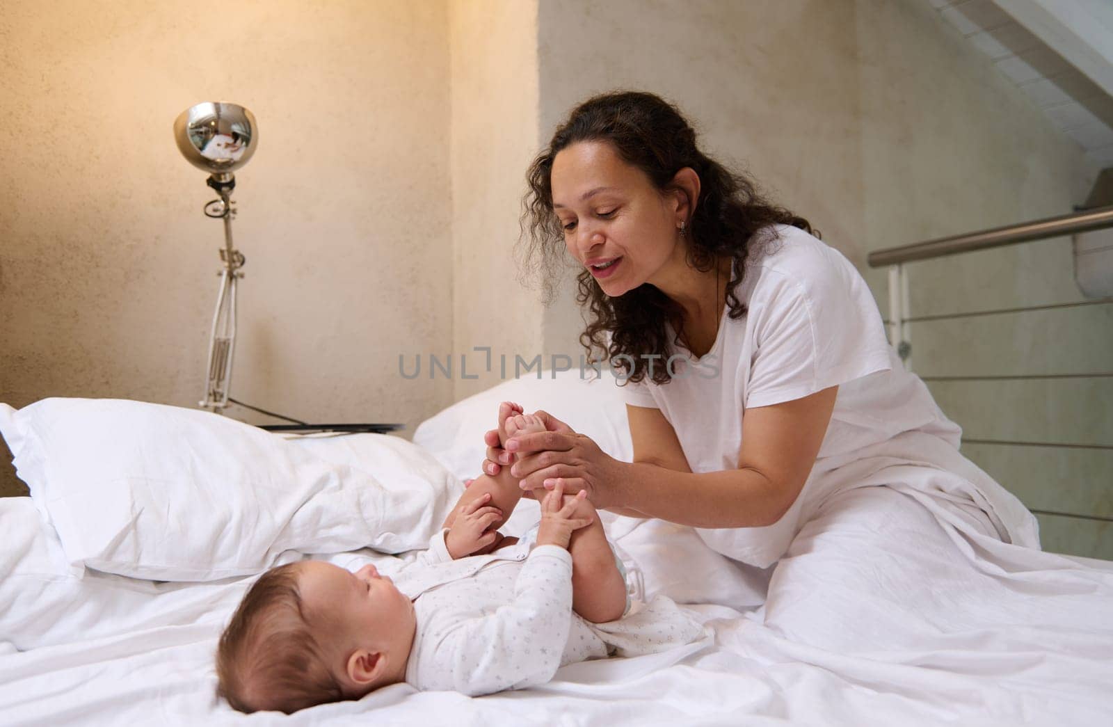 Happy delightful affectionate young mother caring of her newborn baby boy, stroking and kissing his little feet and toes by artgf
