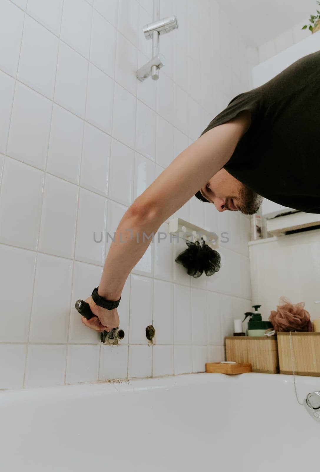 One young Caucasian recognizable man cleans with a screwdriver a hole in a tiled wall with a faucet tube in the bathroom, standing at an angle, close-up side view.Step by step.