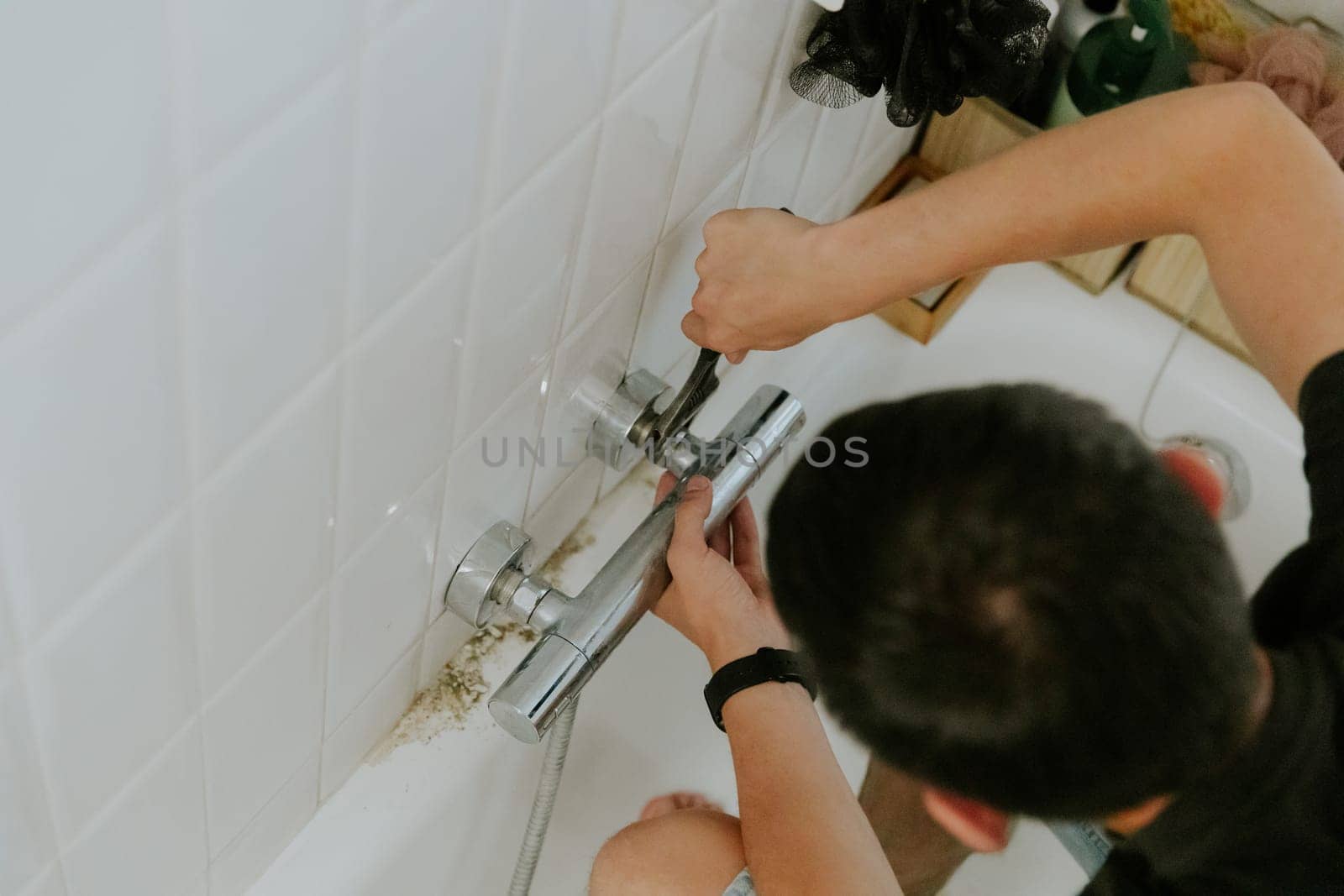 One young Caucasian brunette man manually tightens the nut on the faucet in the wall using an adjustable wrench while sitting on the edge of the bathtub, top view. Step by step.