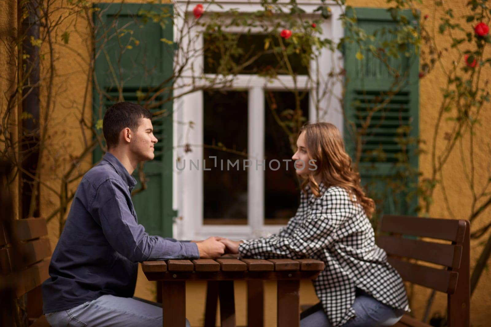 Teen Sweethearts: Holding Hands at an Outdoor Café in a European Town. Happy smiling young dating couple having coffee together and enjoying life sitting at table holding hands in street cafe on summer day. Pretty man and woman spending time together enjoying weekend. Dating, young fashion elegant stylish couple sitting at a table in a cafe , gently hold hands, on streets of old european city. American beautiful man hugging caucasian woman and sitting at cafe, hat on table. Concept of happy international c by Andrii_Ko