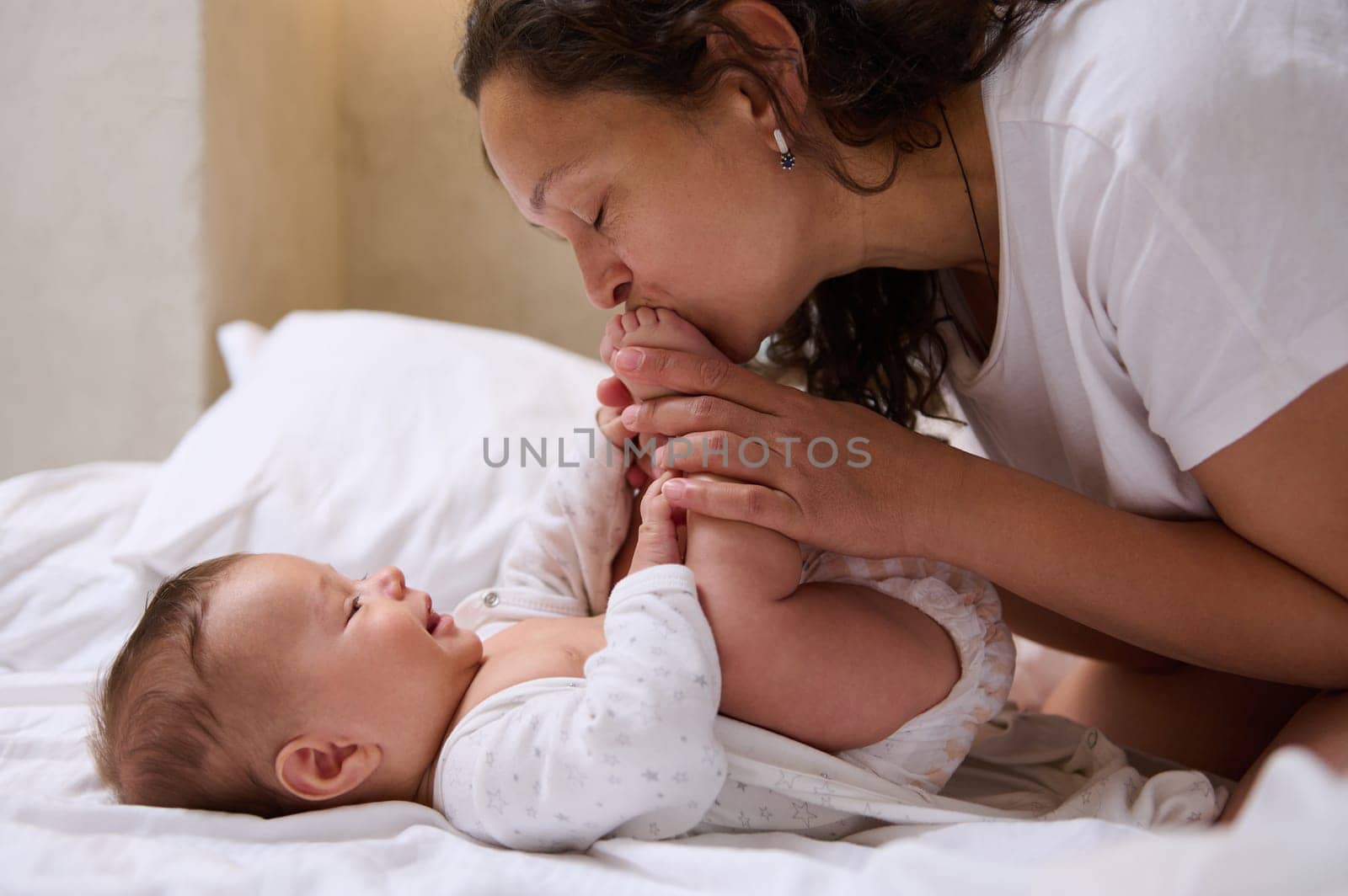 Close-up family of a young adult multi ethnic mother and cute baby boy enjoying spending happy moments together. Mom kissing her baby feet and toes, stroking and embracing him with tenderness and love