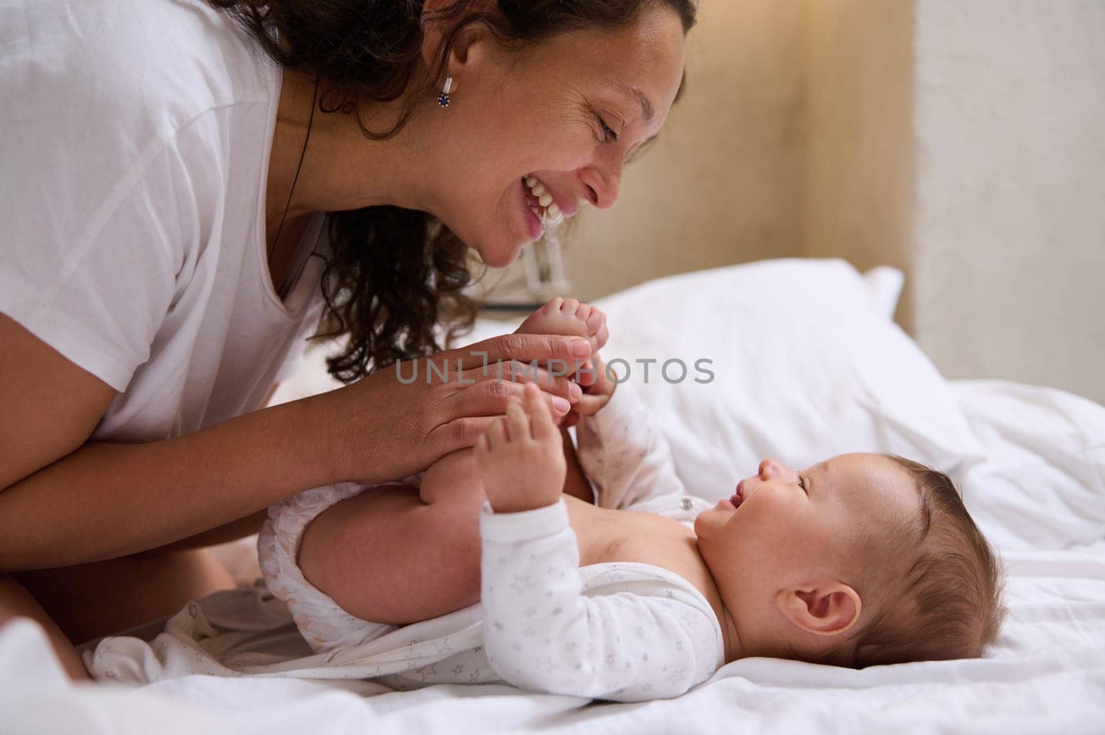 Close up portrait of happy young adult woman, a loving caring mother holding legs of her newborn baby near her face, smiling and looking tenderly at him while kissing him and spending time in bedroom