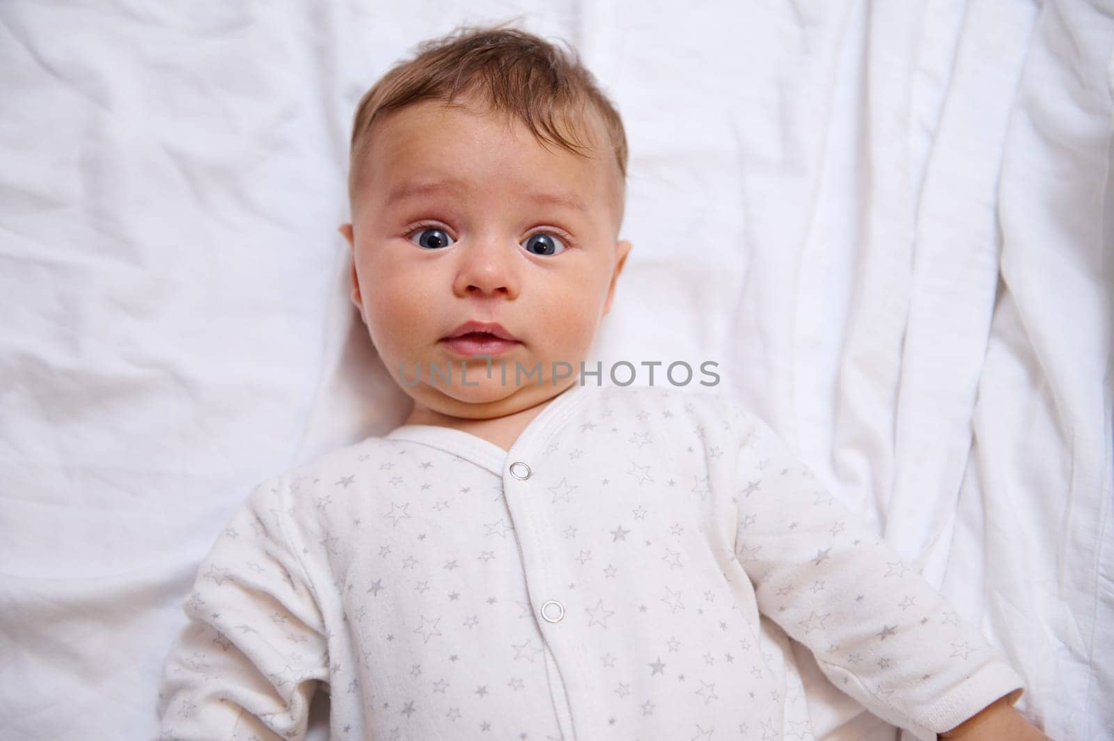 Close-up face portrait of Caucasian adorable lovely beautiful cute baby boy 4-6 months old looking at camera, lying on white bed sheets on the bed. Copy advertising space. Beautiful kids. Baby care
