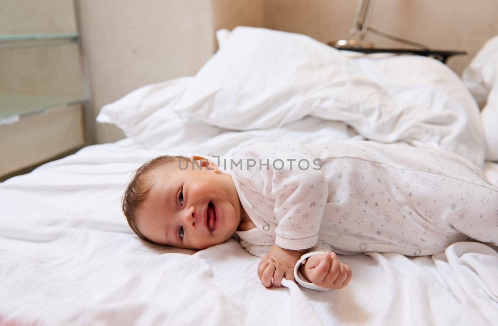 Playful lovely baby boy in white clothes, smiling and laughing lying on white bedsheets on the bed at home by artgf
