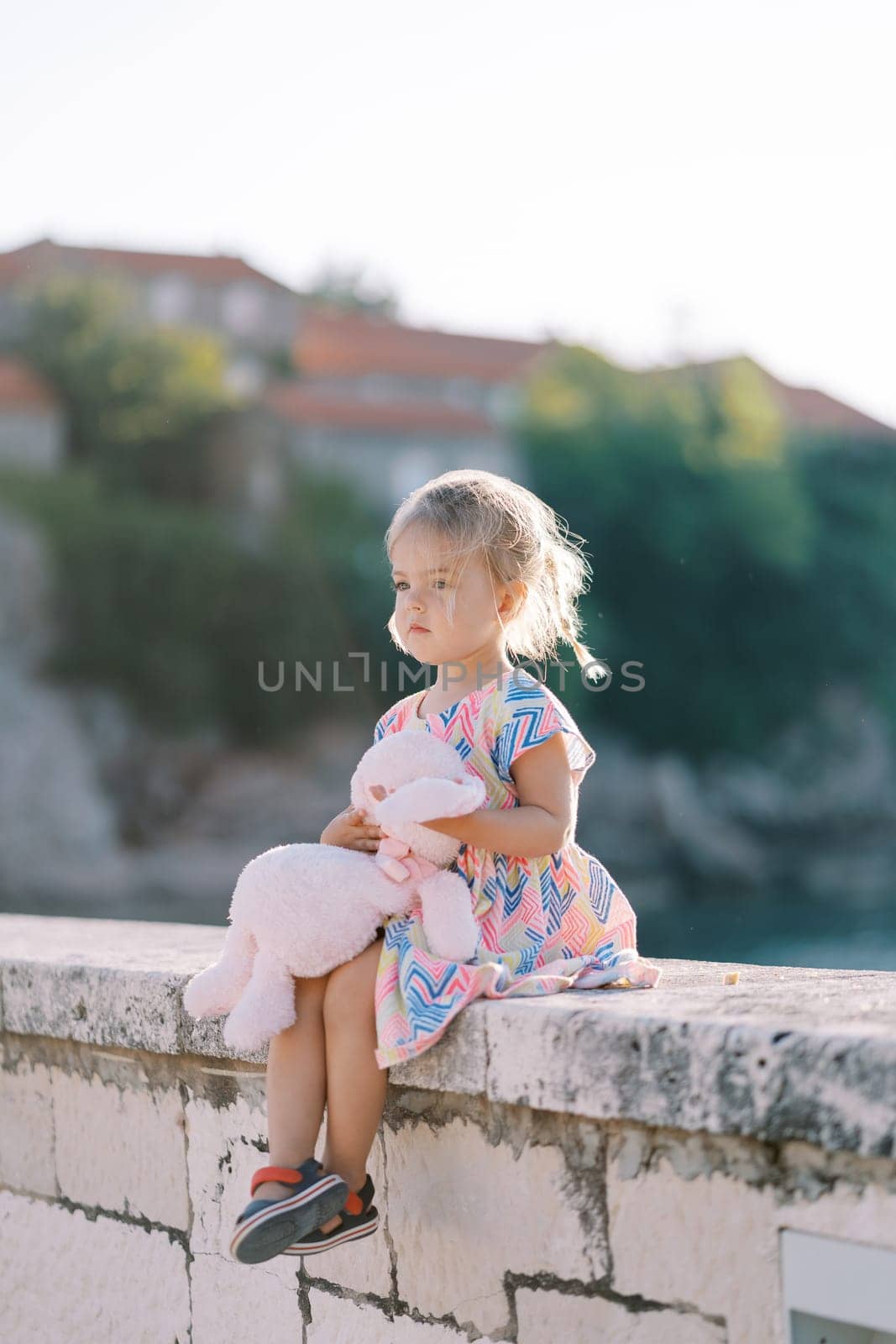 Little girl with a soft toy sits on a stone fence and looks ahead by Nadtochiy