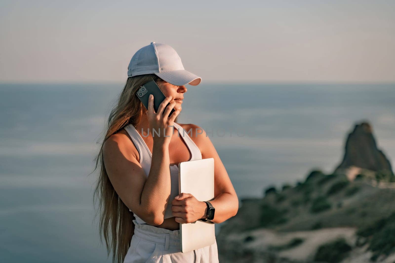 Freelance women sea working laptopr. Happy woman talking on the phone outdoors with a beautiful view of the sea in her hand with a laptop. Remote work concept. by Matiunina