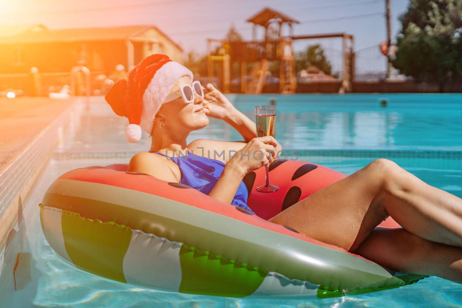 A happy woman in a blue bikini, a red and white Santa hat and sunglasses poses in the pool in an inflatable circle with a watermelon pattern, holding a glass of champagne in her hands. Christmas holidays concept. by Matiunina