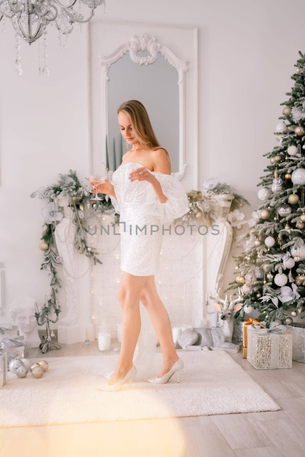 The blonde in the Christmas room. A beautiful blonde woman in a shiny light short dress with a train stands in a beautiful bright room decorated with a festive interior with a Christmas tree. by Matiunina