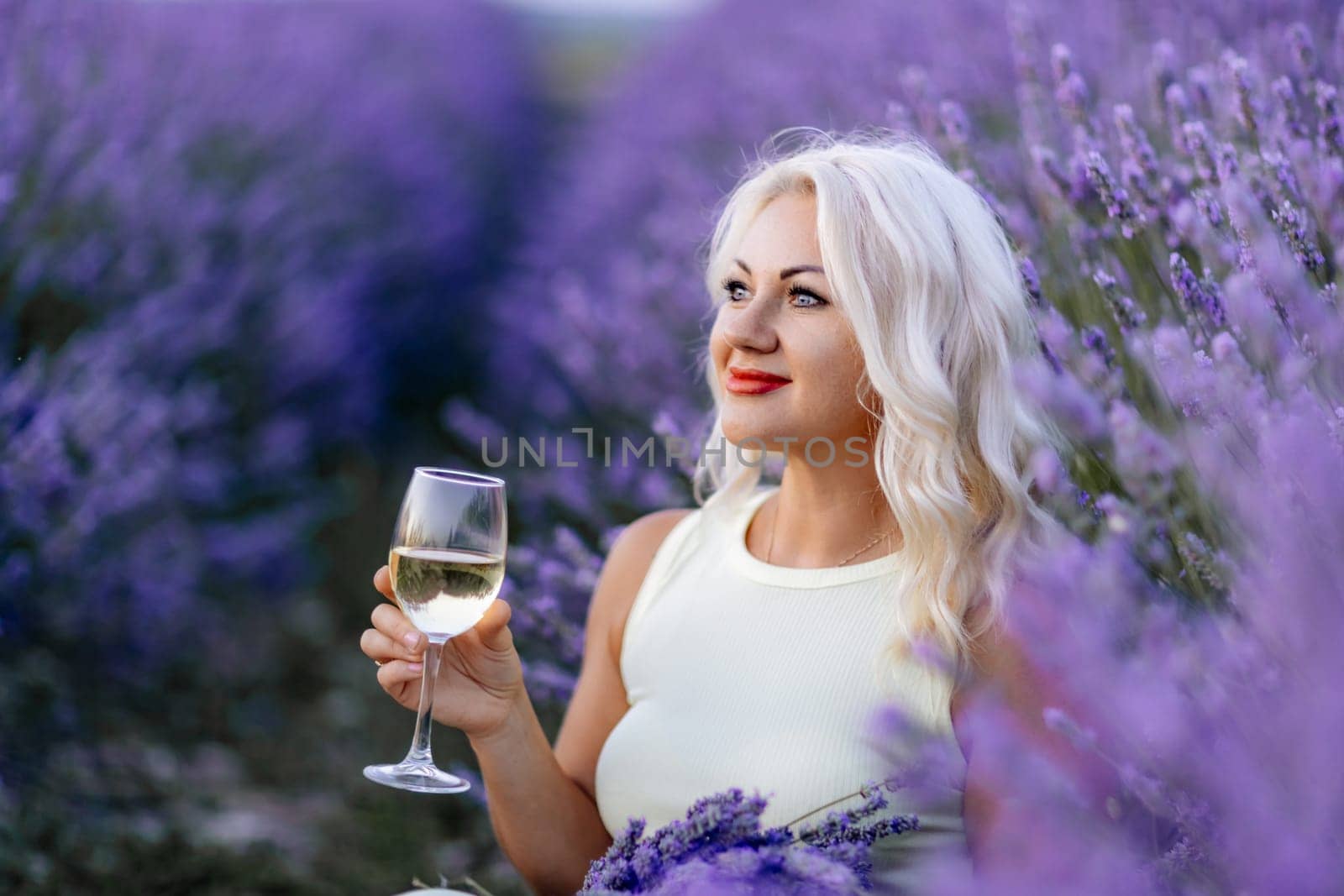 Blonde lavender field holds a glass of white wine in her hands. Happy woman in white dress enjoys lavender field picnic holding a large bouquet of lavender in her hands . Illustrating woman's picnic in a lavender field