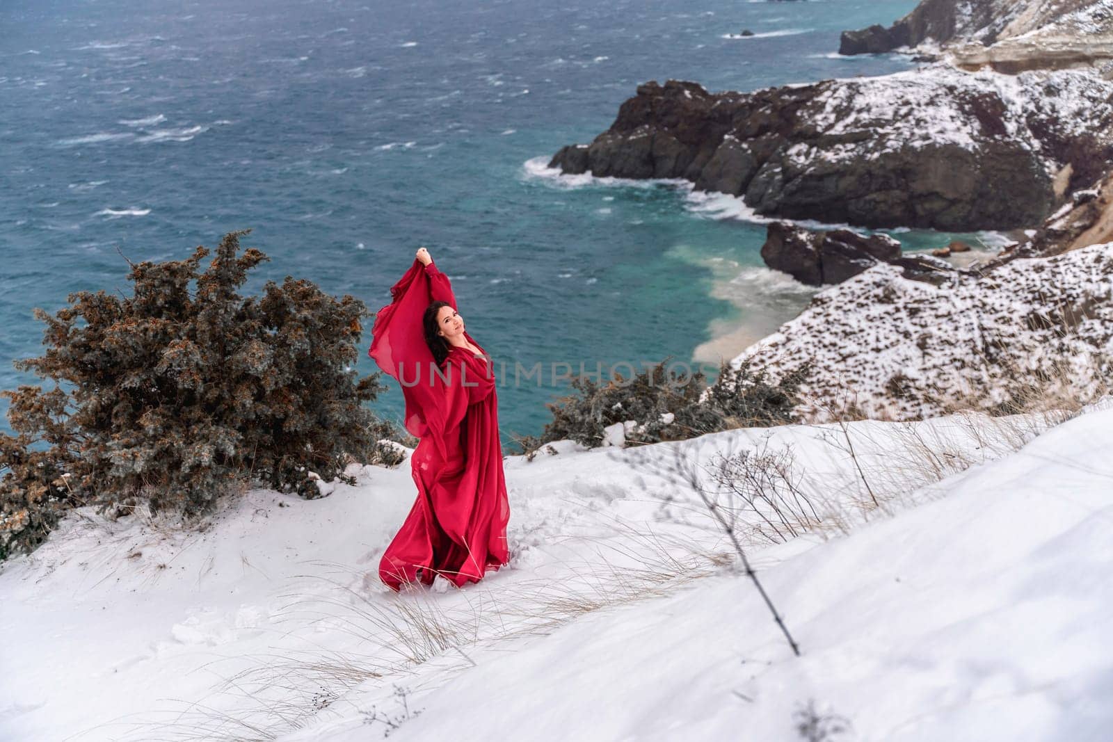 Woman red dress snow sea. Happy woman in a red dress in the snowy mountains by the emerald sea. The wind blows her clothes, posing against sea and snow background. by Matiunina