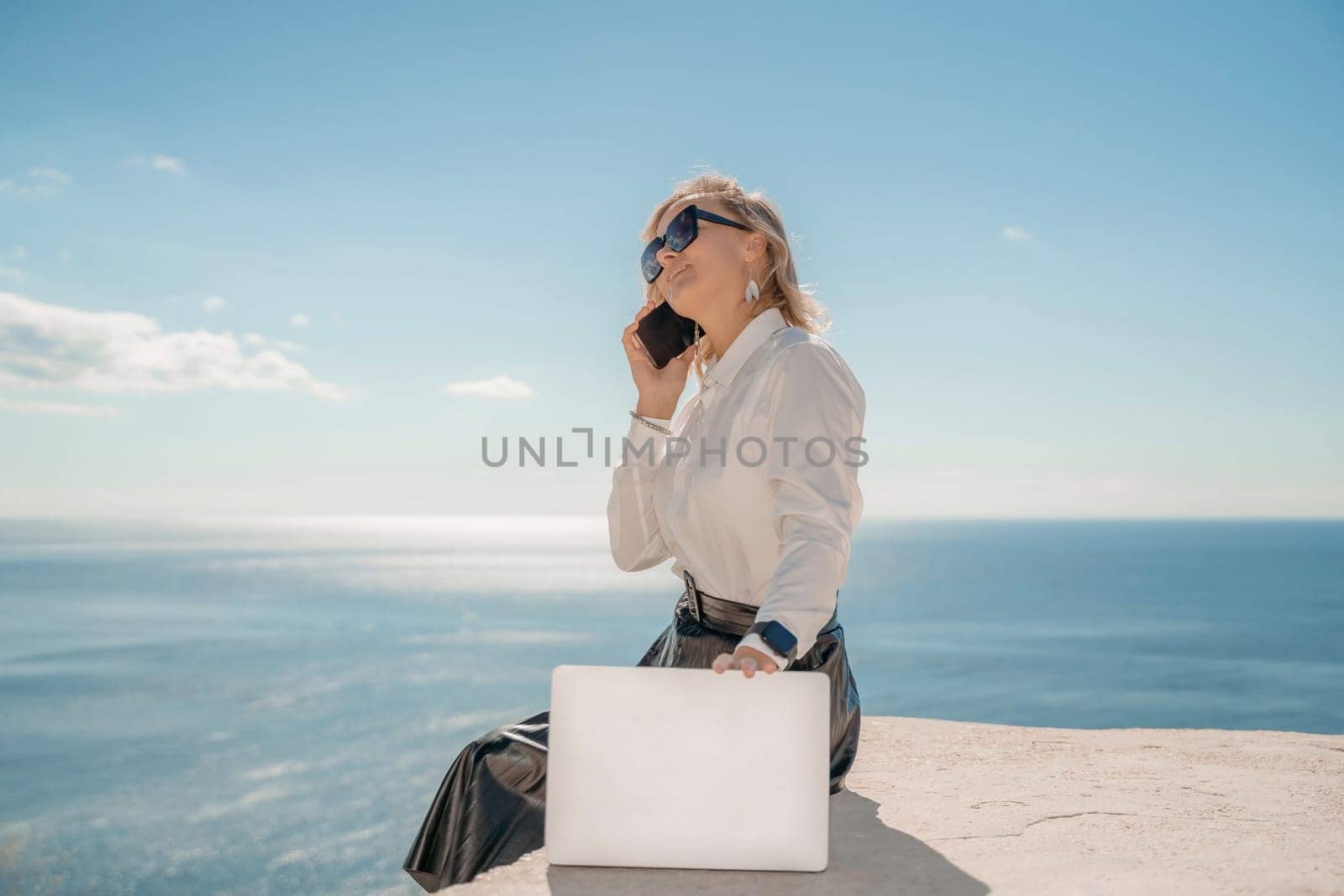 Freelance women sea working on a computer. Pretty middle aged woman with computer and phone outdoors with beautiful sea view. The concept of remote work. by Matiunina