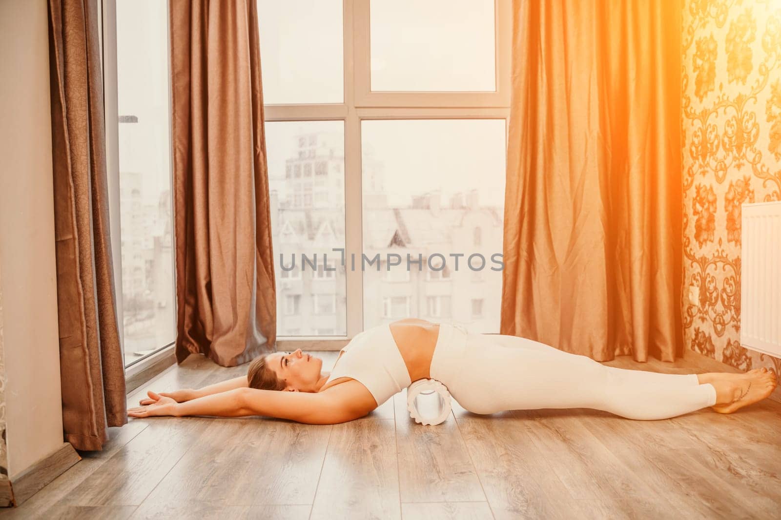Adult athletic woman, in white bodysuit, performing fascia exercises on the floor - caucasian woman using a massage foam roller - a tool to relieve tension in the back and relieve muscle pain - the concept of physiotherapy and stretching training.