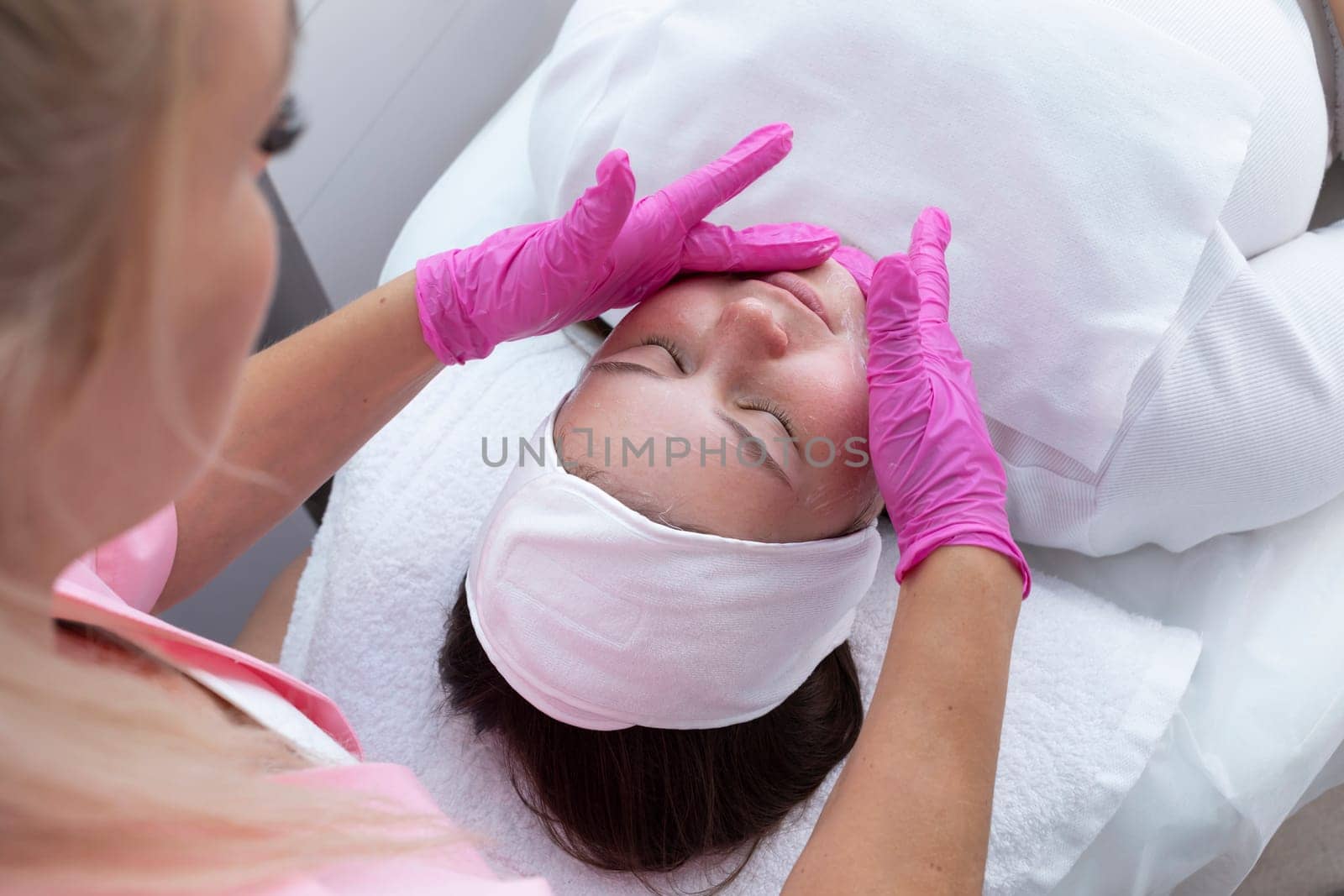 Top View Of Beautiful Young Caucasian Girl During Procedure Of Cleansing, Moisturizing Face Skin, Facial Massage. Professional Female Beautician in Beauty Salon. Horizontal. High quality Photo