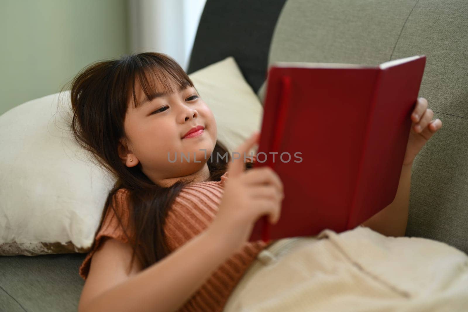 Cute little girl reading book, enjoying interesting story while lying on couch at home