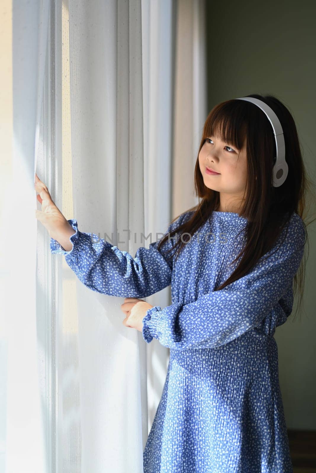 Portrait of pleased Asian child girl listening to music in headphones and looking away through window
