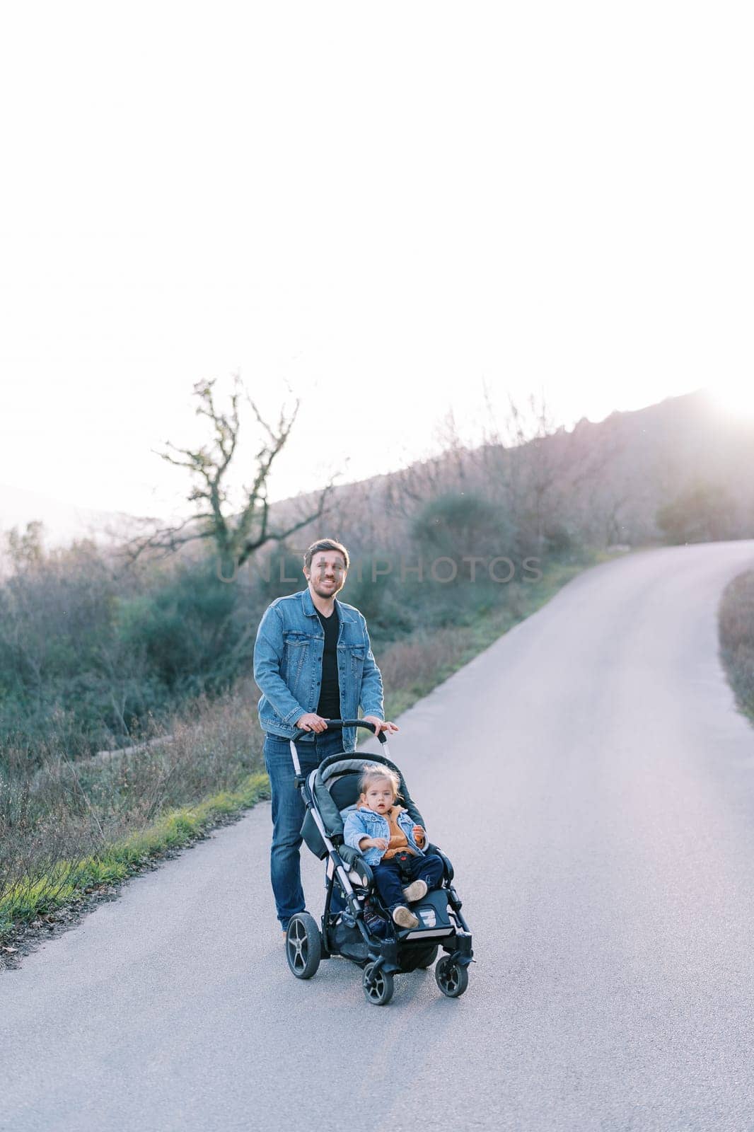 Smiling dad with a little girl in a stroller stands on an asphalt road in the mountains by Nadtochiy