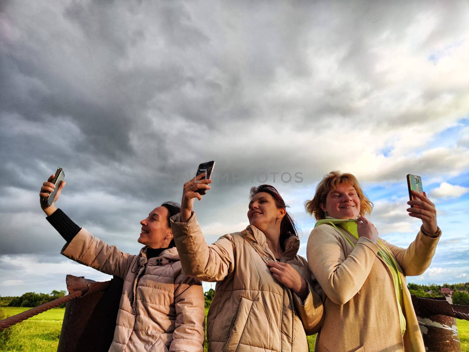 Three funny tourist girls on the old bridge take selfies against the background of an alarming dramatic sky with thunderclouds. Middle-aged women having fun outdoors by keleny