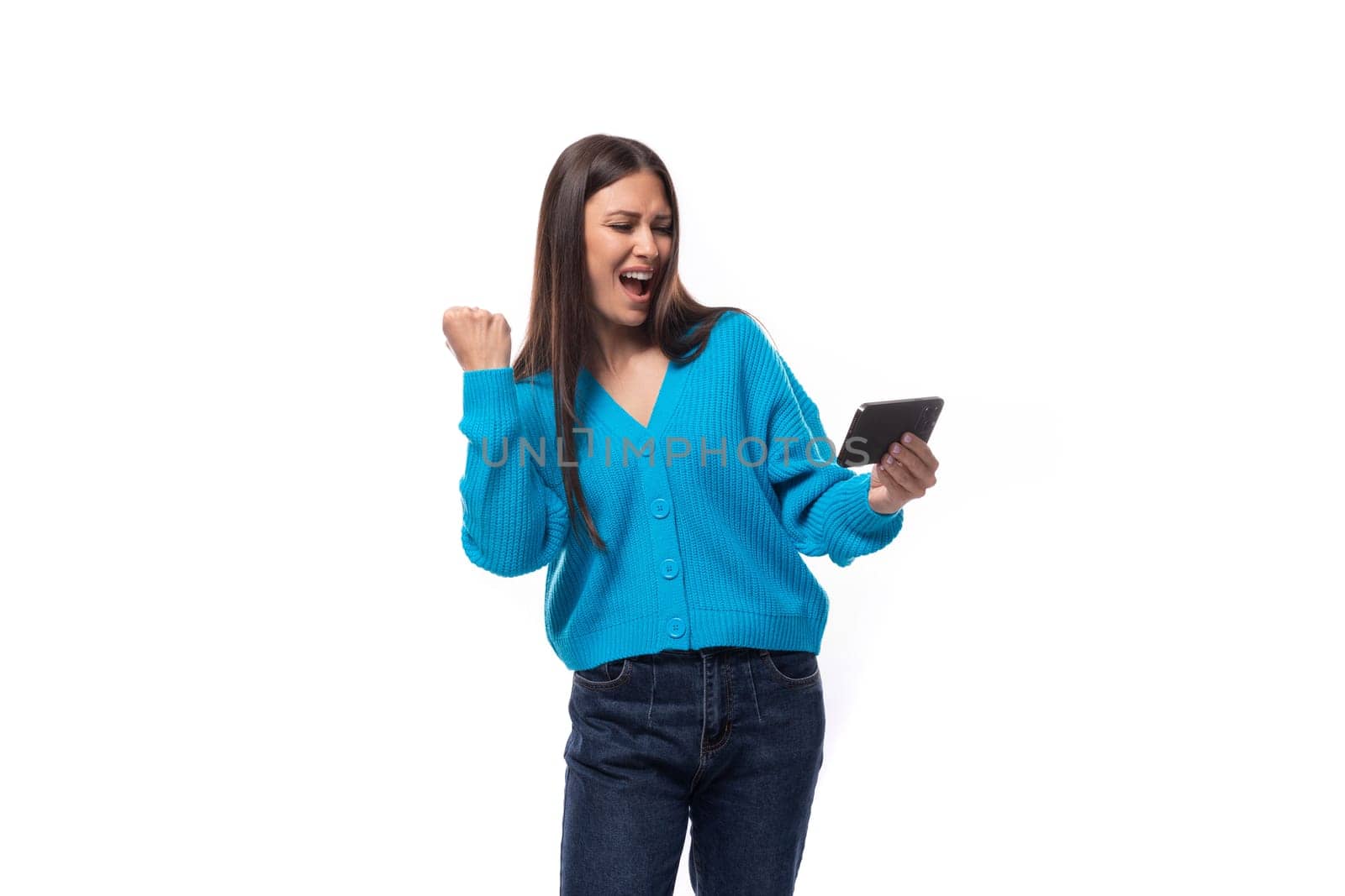 young beautiful european brunette woman dressed in blue cardigan rejoices watching video on phone.