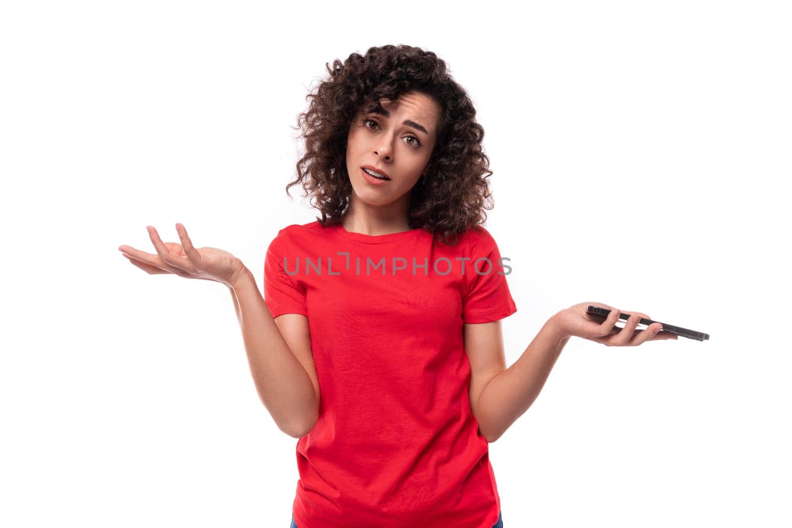 caucasian young pretty curly brunette woman in a red t-shirt spreads her arms in excitement holding a smartphone in her hand by TRMK