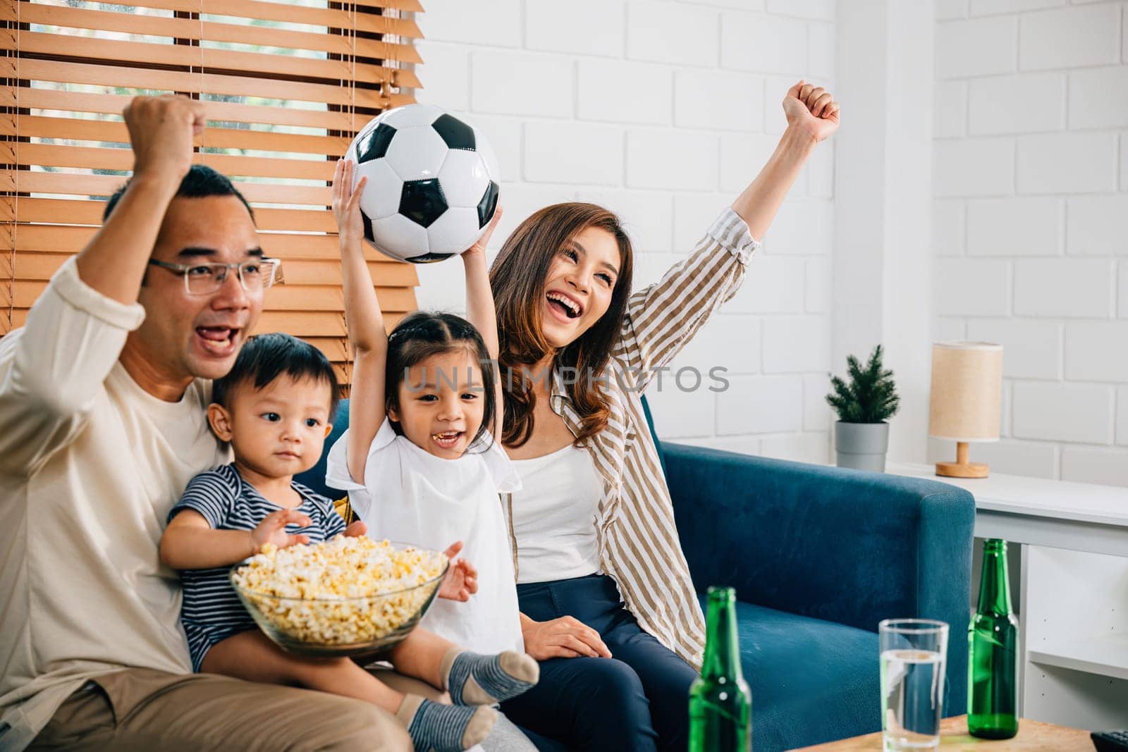 Delighted parents and children enjoy a football match at home, celebrating a goal with popcorn and a ball. Their excitement and togetherness capture the joy of winning and family fun.