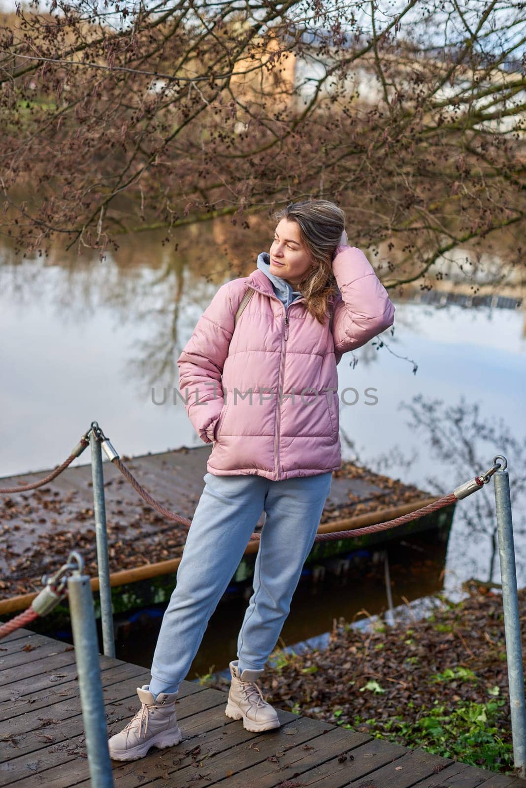 A young woman standing at the shore looking at the river in autumn sunny day. Street view, copy space for text, travel photo. Happy tourist woman on the bank of the river in autumn in warm clothes. Tourists enjoy their vacation, winter season. Romantic look and travel concept. A joyful mood in a caucasian girl. Winter Wonderland: Enchanting Girl by the Riverside in Autumn by Andrii_Ko